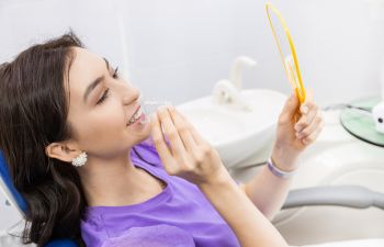 Happy woman holding clear dental aligner looking at her nice smile in a mirror.